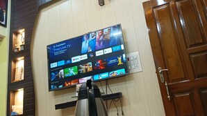 A room with a flat-screen TV mounted on the wall displaying various streaming services. There is a stainless steel kettle on a table with a floral tablecloth. The room has wooden paneling on the walls and a wooden door. - Trilok Residency  (Varanasi)