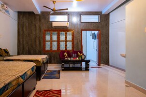 A hotel room with two beds, a ceiling fan, an air conditioning unit, a window with a decorative grille, a small table with a vase and a kettle, and a door with a security camera.