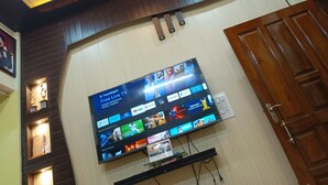 A flat-screen TV mounted on a wall with a wooden panel design, displaying various streaming apps and a "PatchWall+ Free Live TV" interface. - Trilok Residency  (Varanasi)