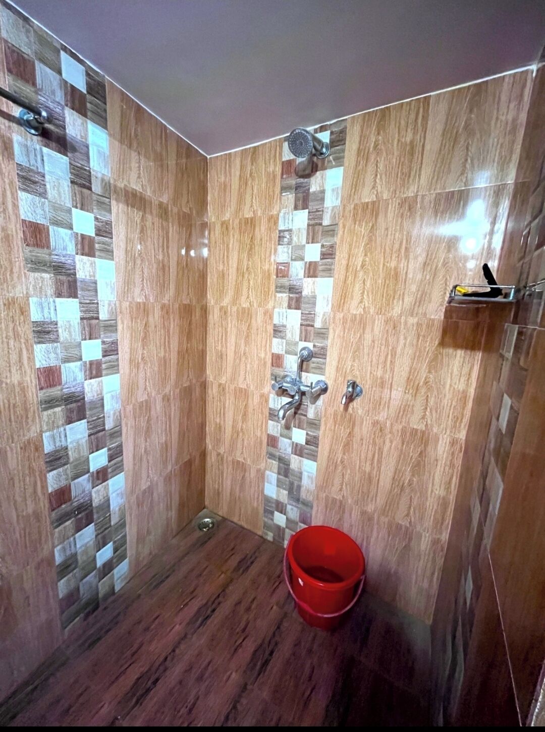 A shower with a checkered tile pattern, a red bucket, and a metal shelf.