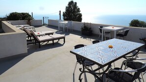 A rooftop terrace with lounge chairs, a table, and chairs, overlooking a scenic view of the sea.