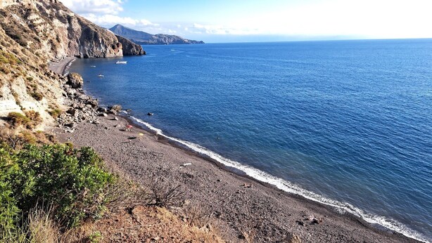 A coastal landscape with a rocky beach, clear blue sea, and distant mountains.