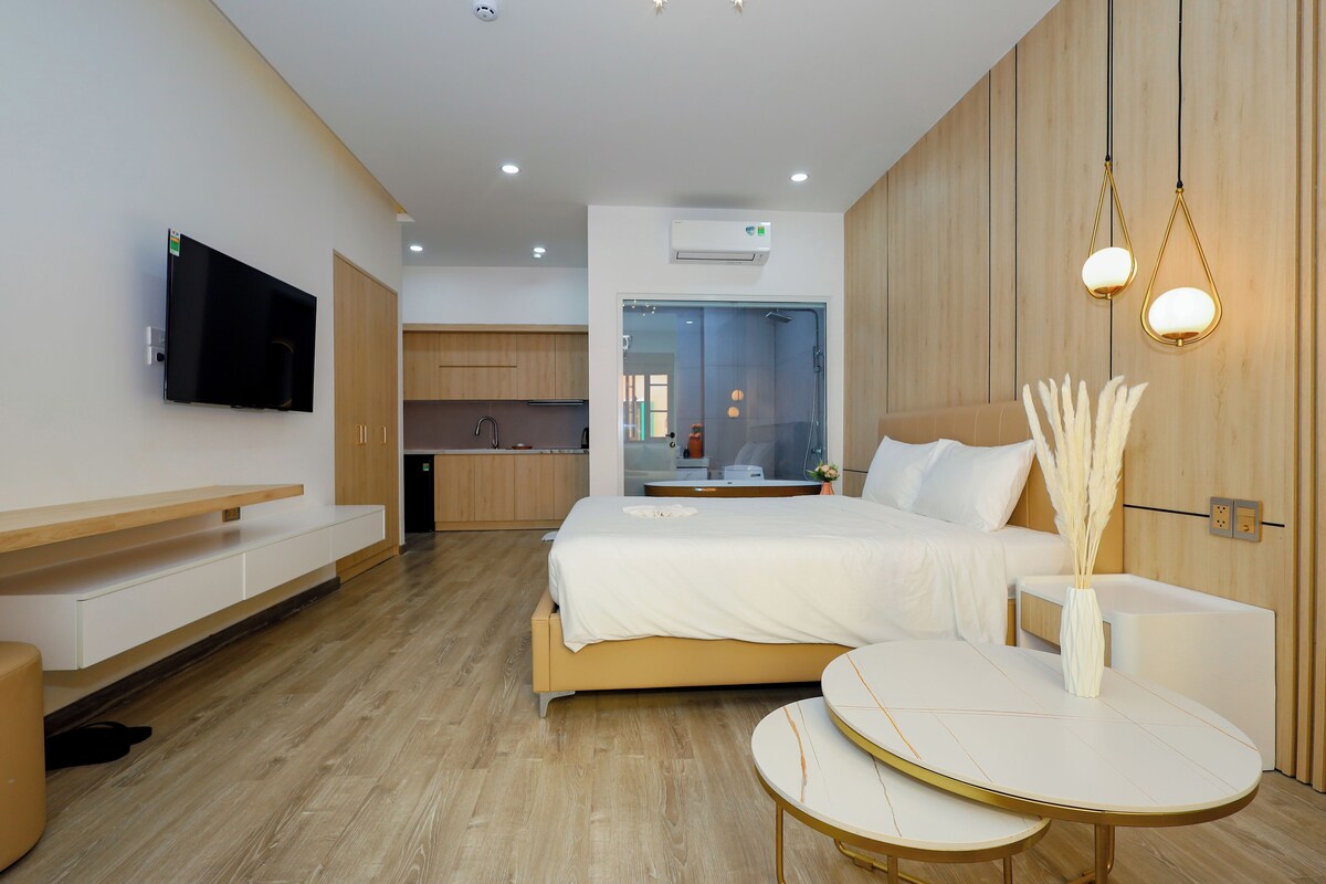 Ein modernes Hotelzimmer mit einem Bett, einem kleinen Tisch, einem Fernseher und einer Küchenzeile im Hintergrund.