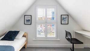 A bedroom with a sloped ceiling, a bed with a blue blanket, a desk with a chair, and a window with blinds.