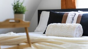 A neatly made bed with white and dark pillows, a white throw blanket, and a wooden nightstand with a plant.