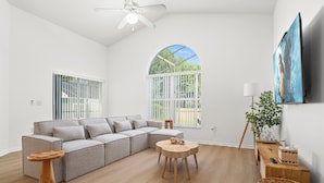 A modern living room with a sectional sofa, a round wooden coffee table, a small side table, a TV mounted on the wall, and a large window with a view of trees. - Highgate@Legacy Park, a 3 bed 2 bath pet friendly home within a gated community (Davenport)