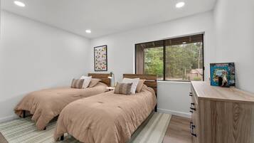 Un dormitorio con dos camas, una ventana con vista a árboles, una mesita de noche con una lámpara y una estantería con un libro.