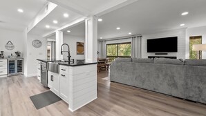 A modern kitchen with white cabinetry, a central island, and a dining area with a gray sofa and a flat-screen TV. - COMPLETELY REMODELED 3 BDR, 2 Bath, Golf Course View Condo (Pinehurst)