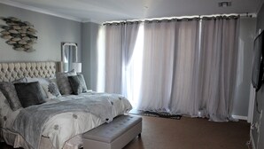 A bedroom with a tufted headboard, a large bed with grey bedding, a bench, a wall-mounted TV, and sheer curtains.