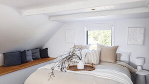 A bedroom with a sloped ceiling, a wooden bench along the wall, a bed with multiple pillows, a bedside table with a lamp, and two framed artworks on the wall.