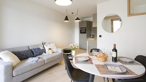 Un salon moderne avec un canapé gris, des chaises noires, une table à manger dressée pour quatre personnes et une cuisine en arrière-plan.