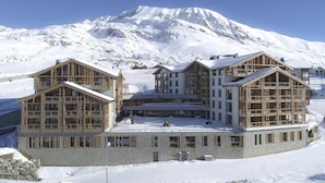 Un chalet à étages, doté d’une architecture symétrique, de grandes fenêtres et de balcons en bois, le tout se dressant sur fond de montagnes enneigées.