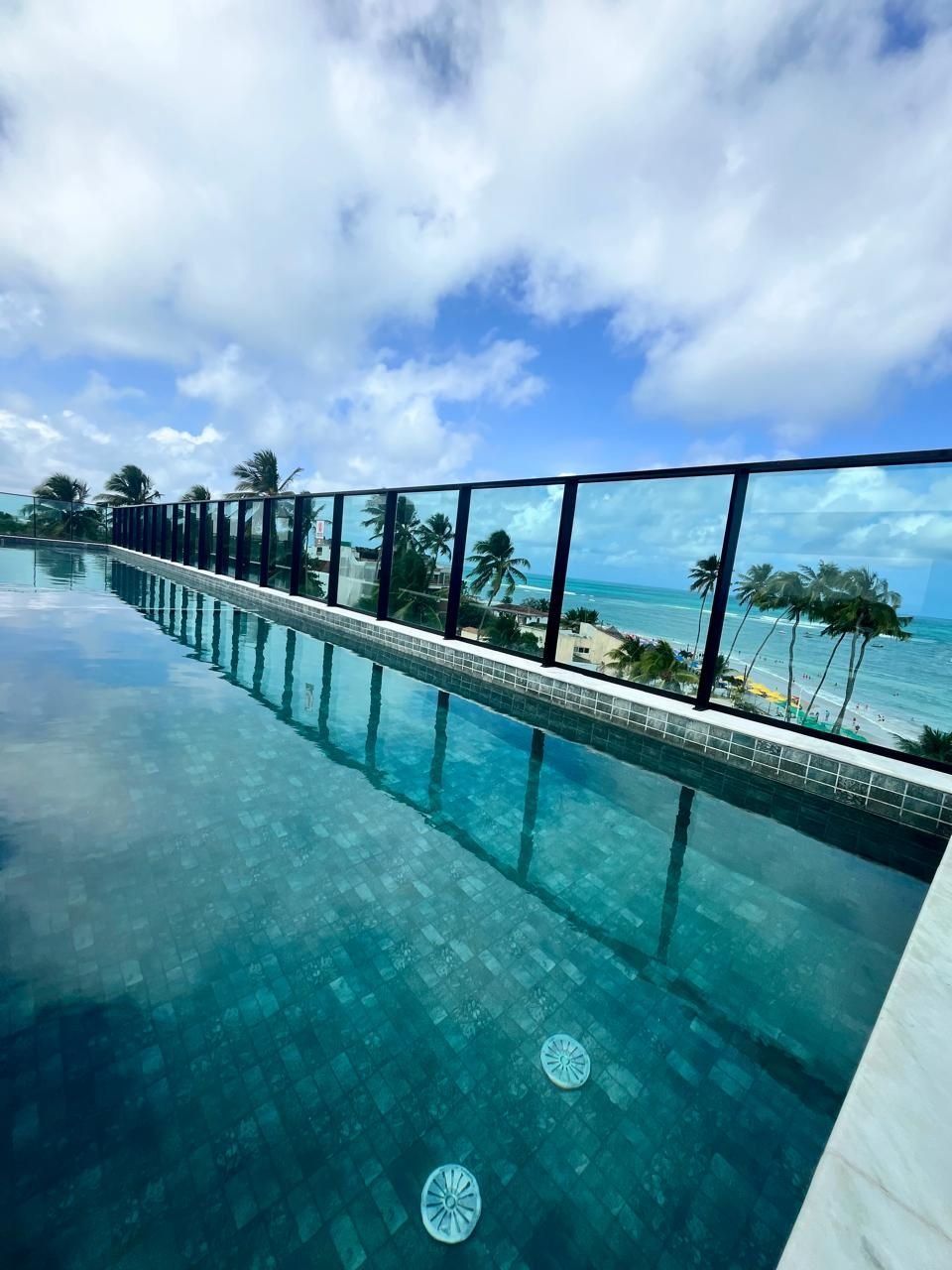 A clear view of an infinity pool with a tiled edge, leading to a beach with palm trees and a clear sky.