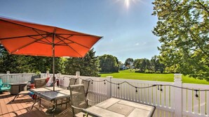 An outdoor deck with a table and chairs, a large red umbrella, and a view of a well-maintained lawn and trees. - Relaxing 5-Bedroom Getaway with Saltwater Pool & Game Room Close to Hershey (Palmyra)