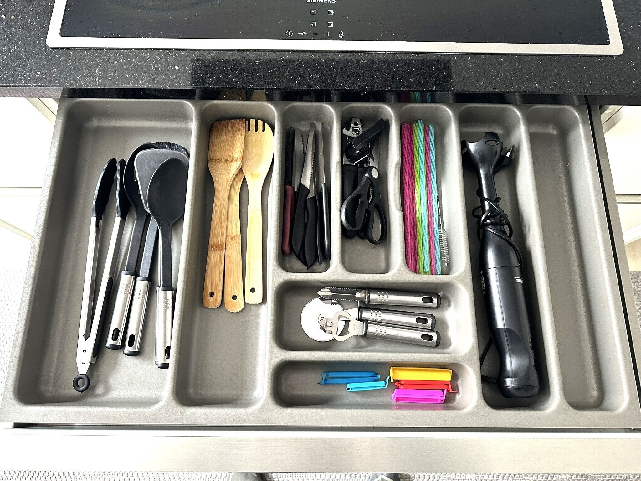 Uma gaveta de cozinha com diversos utensílios e ferramentas, incluindo facas, espátulas e um processador de alimentos.