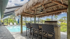 An outdoor pool area with a thatched roof, bar with chairs, and a view of the surrounding landscape.