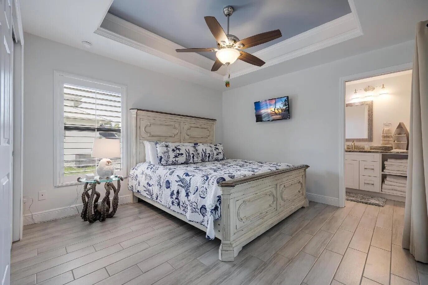A bedroom with a bed, a ceiling fan, a window with blinds, a painting on the wall, and a bathroom visible through an open door.