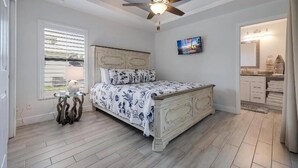 A bedroom with a bed, a ceiling fan, a window with blinds, a painting on the wall, and a bathroom visible through an open door.
