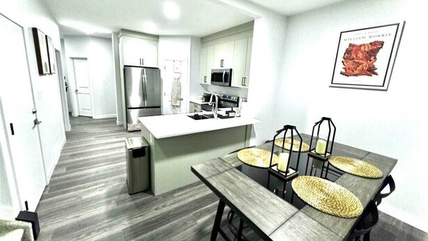 A modern kitchen with white cabinets, a stainless steel refrigerator, and a microwave. There is a dining area with a wooden table and woven placemats. - Modern Marvel in Windermere (Edmonton)