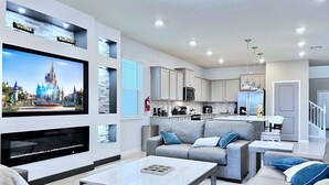 Una sala de estar moderna con una televisión de pantalla plana, sofás grises, una mesa de centro y una zona de cocina con armarios blancos y electrodomésticos de acero inoxidable.