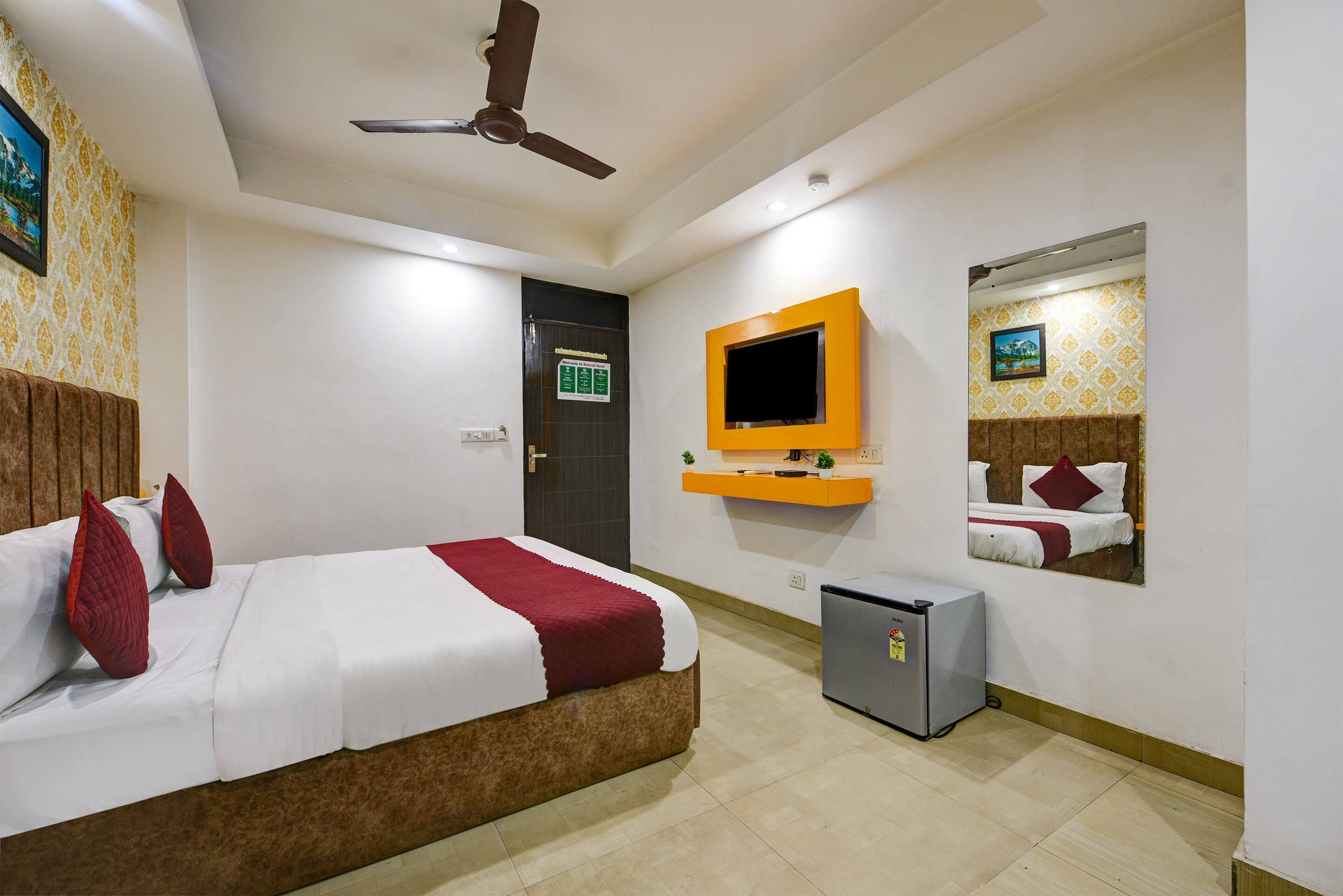 A hotel room with a bed, a ceiling fan, a TV mounted on the wall, a small fridge, and a door with a sign.