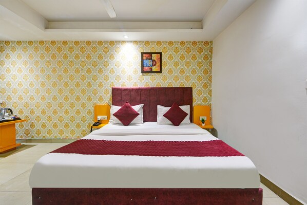 A hotel room with a large bed, two bedside tables, a nightstand, a wall-mounted picture, and patterned wallpaper. - Hotel Happy Stay - Delhi Airport (New Delhi)