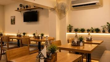 A restaurant interior with wooden tables and chairs, a flat-screen TV mounted on the wall, and an air conditioning unit.