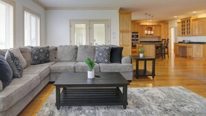 A living room with a sectional sofa, a coffee table, and a small side table with a plant. There is a kitchen area with wooden cabinets and a dining area visible in the background. - "Endless Summer" Waterfront on the Chesapeake Bay! (Woolford)