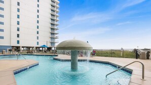 A swimming pool with a central fountain, surrounded by a fence, and adjacent to a multi-story building. - Phoenix 8 8904- Sand Shells Sun Can Rent at 18 3 Bedrooms 2 Baths (Orange Beach)
