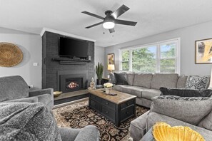 Un salon avec une cheminée, des canapés gris, une table basse et un ventilateur de plafond.