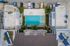 Vista aérea de una piscina en la azotea rodeada de edificios, con paneles solares y coches estacionados.