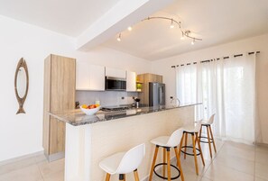 Una cocina moderna con encimera de mármol, armarios de madera y horno empotrado. Hay dos taburetes blancos para bar y un cuenco con fruta sobre la encimera.
