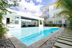Ett modernt poolområde med en klarblå pool, en täckt utomhusplats med sittmöbler och en tvåvåningsbyggnad med balkonger.