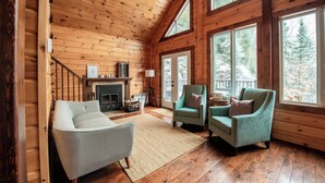A cozy cabin interior with a fireplace, two armchairs, and large windows offering a view of the forest.
