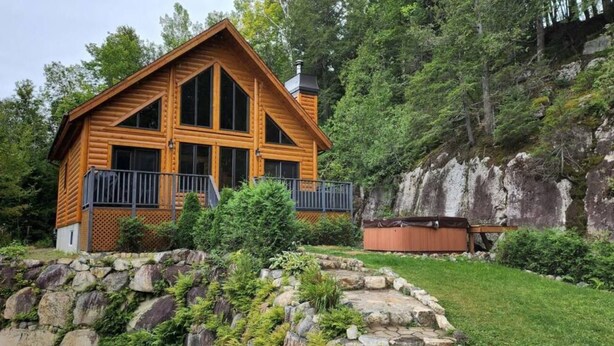 A log cabin with a balcony, surrounded by a stone wall and lush greenery.