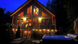 A wooden cabin with a balcony, a hot tub, and a staircase leading to the entrance.