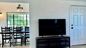 Un salón amplio con una televisión grande sobre un soporte negro, un comedor con sillas de madera y una mesa, y una puerta blanca.
