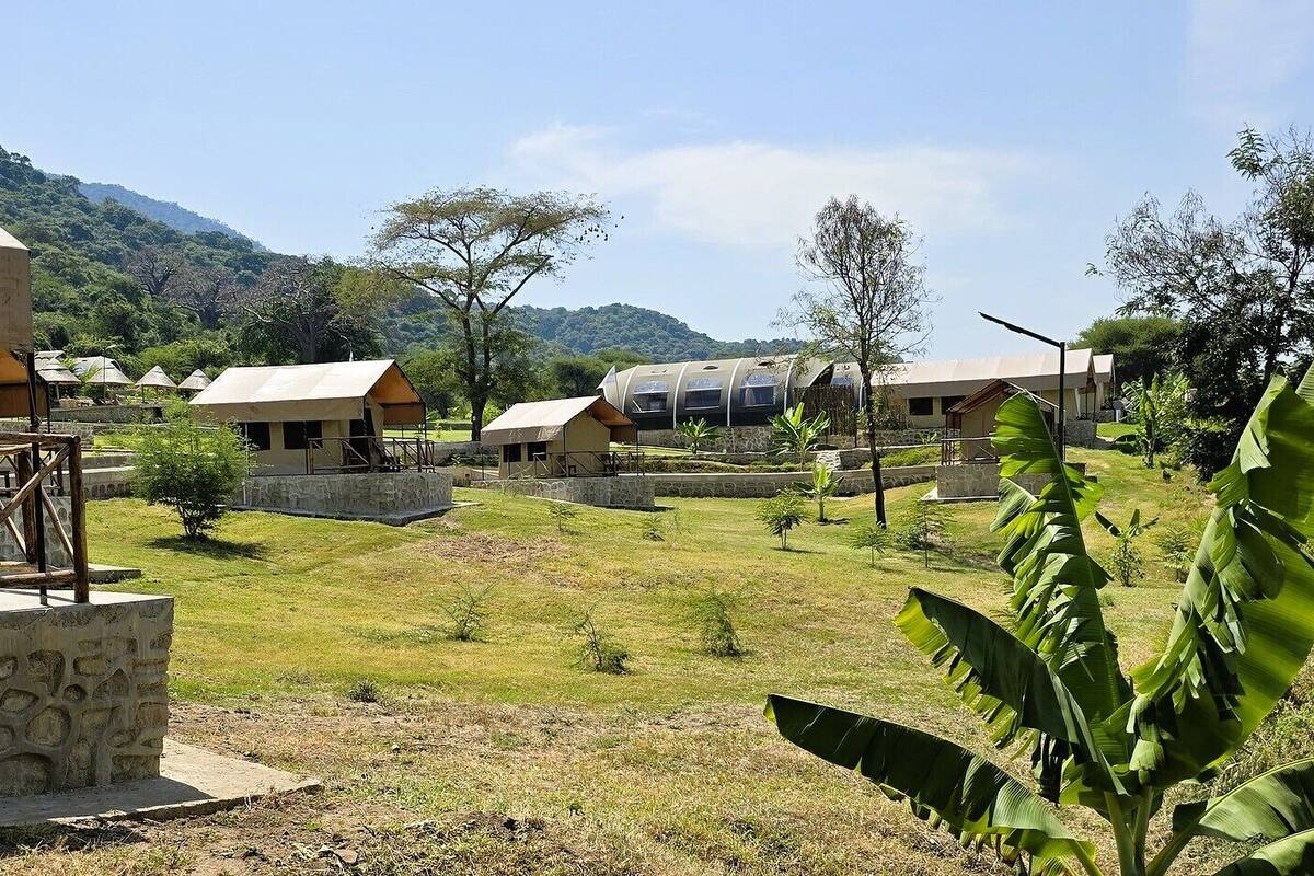 A rural accommodation area with several small, single-story buildings with flat roofs, surrounded by greenery and hills.