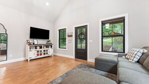 A spacious living room with a flat-screen TV, a white cabinet with storage, a gray sectional sofa, and large windows offering a view of the outdoors. - New Farm Cottage, near Branson, MO built in 2022 (Walnut Shade)