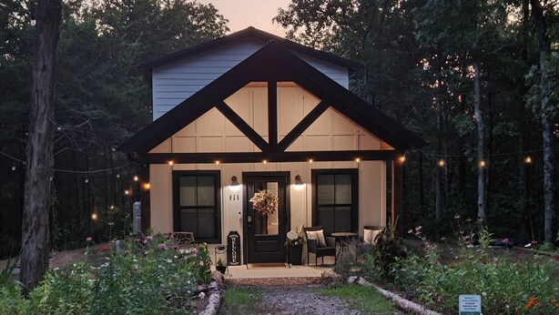 A two-story house with a dark brown roof and white walls, surrounded by a garden with a wooden pathway leading to the front door. - New Farm Cottage, near Branson, MO built in 2022 (Walnut Shade)