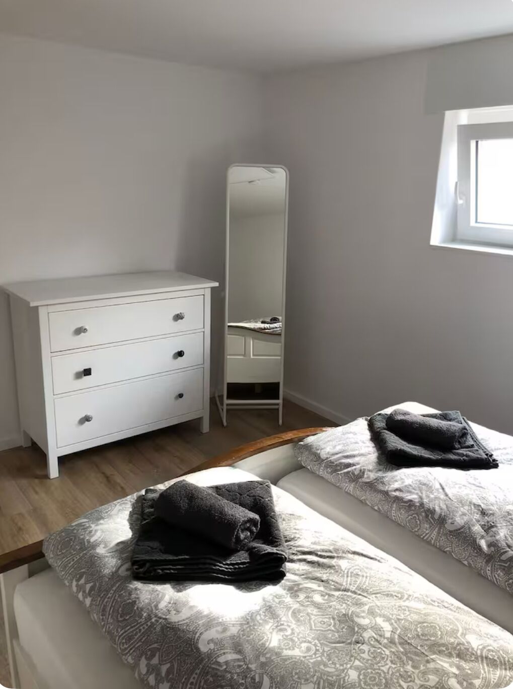 A bedroom with a white dresser, a mirror, two beds with patterned bedding, and a window.