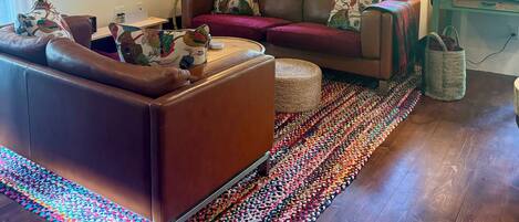 A cozy living room with a brown leather sofa, a colorful woven rug, and a window with a view of greenery.