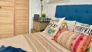 Un dormitorio con cama, cabecera azul, armario de madera, ventana con persianas y mesita de noche con lámpara y objetos decorativos.