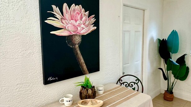 Un comedor con una mesa dispuesta para dos personas, un cuadro de una flor en la pared y una planta en maceta junto a la puerta.
