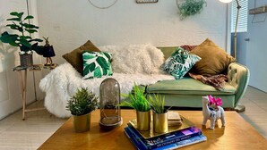 Una sala de estar acogedora con un sofá verde, un plaid blanco mullido y varios objetos decorativos, como plantas y libros.