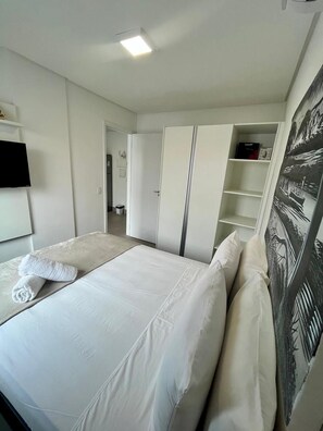 A hotel room with a bed, white linens, a flat-screen TV, a wardrobe, and a painting on the wall. - Green Porto 303 (Ipojuca)