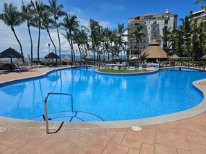 Une piscine à bordures courbes, entourée de palmiers et bordée d’un bâtiment doté de balcons.