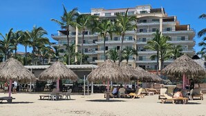 A beach with thatched umbrellas and lounge chairs, a hotel building in the background, and palm trees. - Playa Royale Tower One Studio (Nuevo Vallarta)