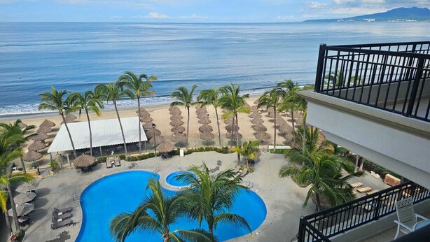 A resort pool area with a view of the ocean, surrounded by palm trees and beach umbrellas. - Playa Royale Tower One Studio (Nuevo Vallarta)