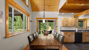 Un grand espace à manger avec une table et des chaises en bois, une grande fenêtre donnant sur un paysage verdoyant, et une cuisine avec des armoires en bois et un évier.
